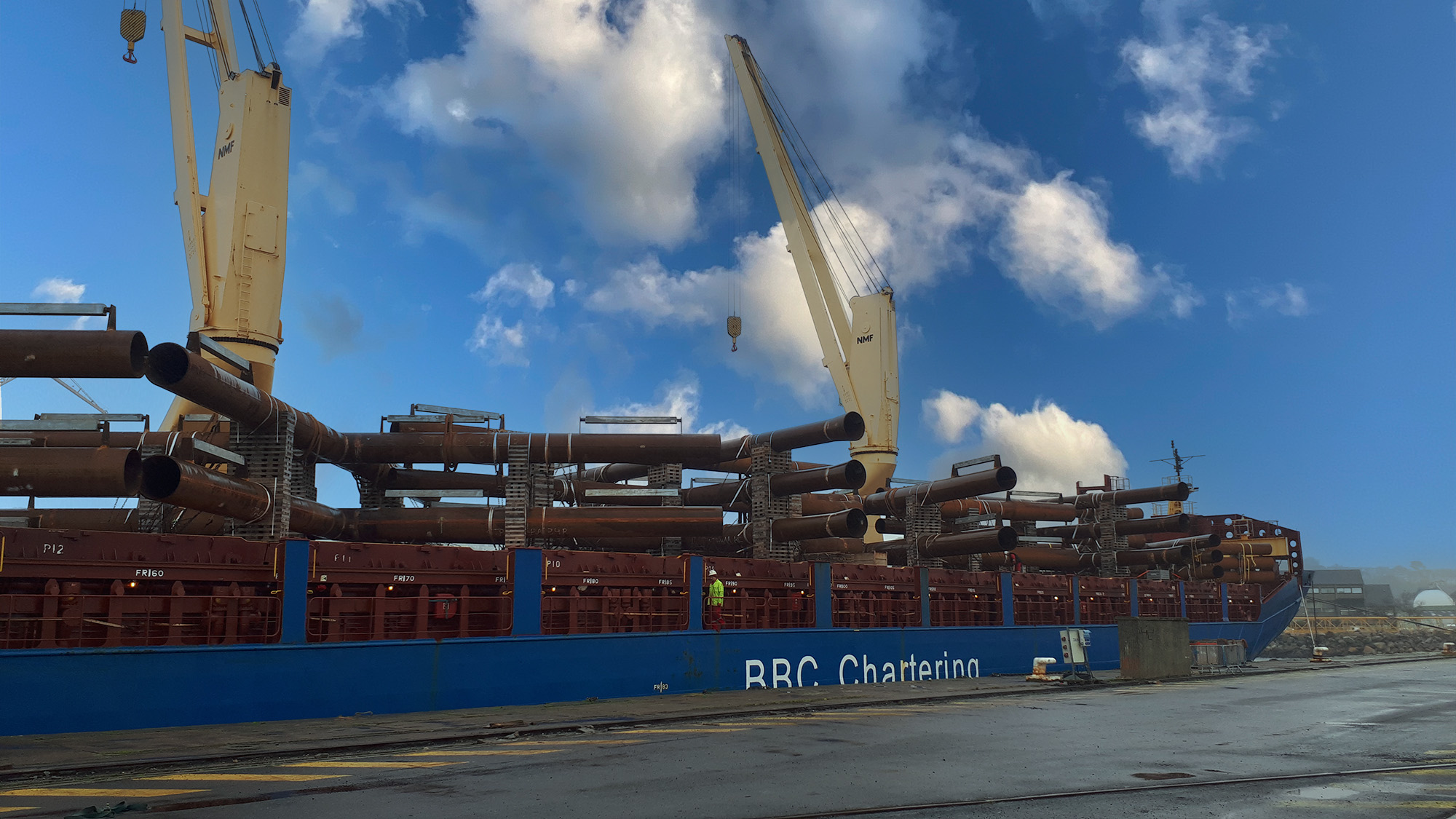 A giant cargo ship docked at a harbor, being loaded with pipes by two cranes.