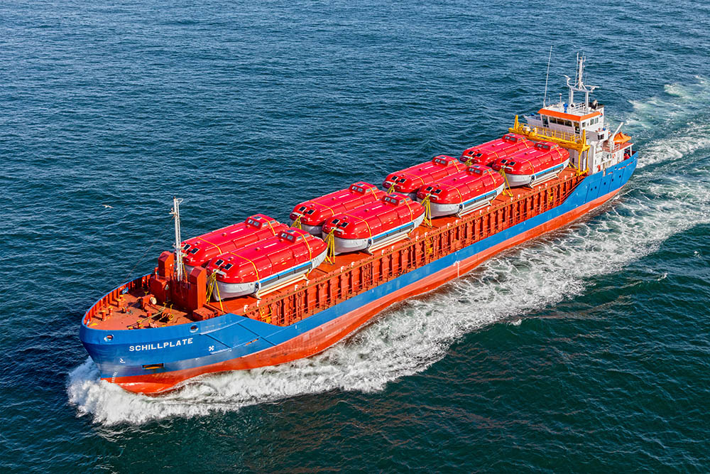 The image shows a large cargo ship carrying orange life rafts on a blue ocean. The text “SCHILLPLATE” is written on the side of the ship, near the bow.
