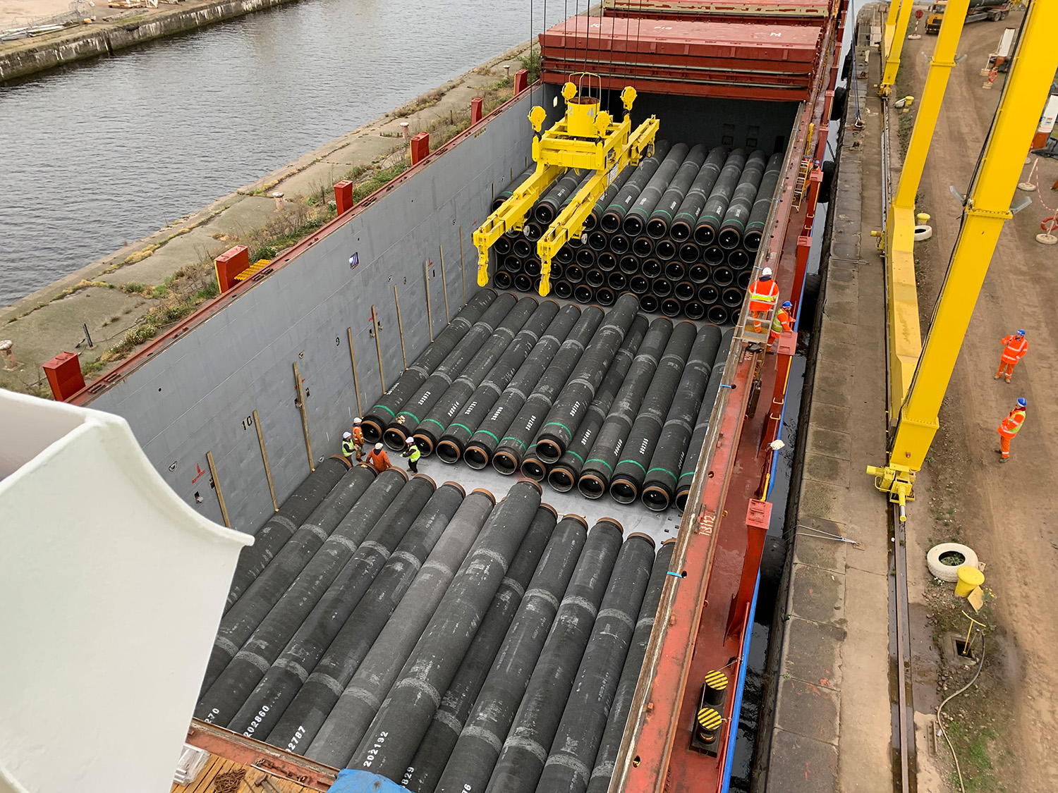 A top view of a cargo ship loaded with black pipes. A crane is lifting one of the pipes.