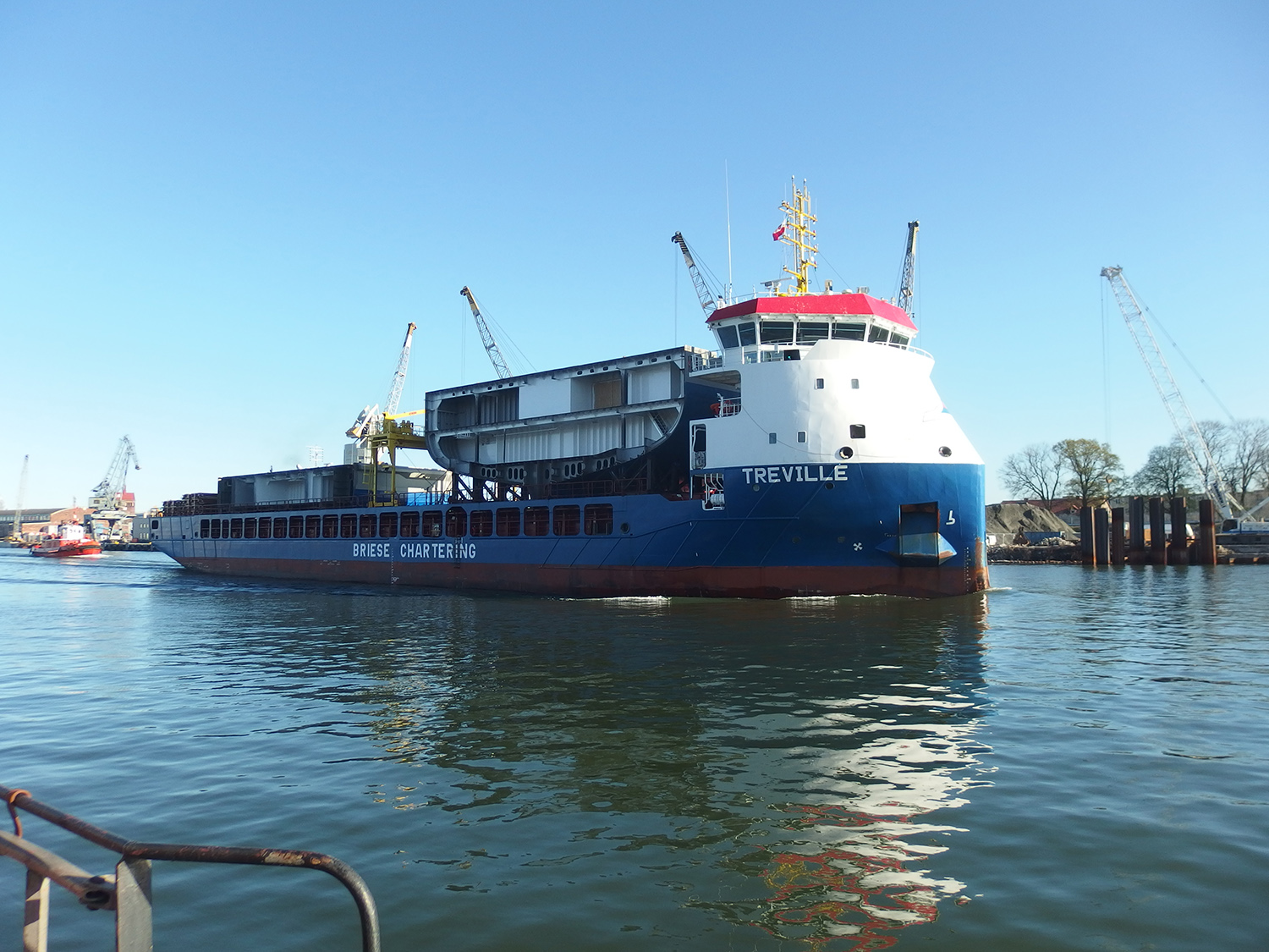 A large blue and white cargo ship named “TREVILLE“ floats on a calm body of water. The text on the ship's side also includes “Briese Chartering GmbH & Co. KG“ and “Mv Aramis General Cargo“. The ship is loaded with 3 shipyard units.