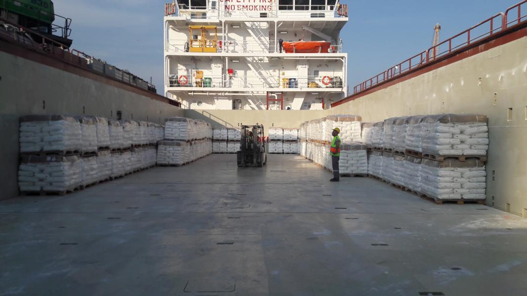 Deck of a cargo ship loaded with bagged goods on pallets, a forklift in the middle, and a worker in a safety vest and helmet overseeing the loading process. The ship's superstructure with safety equipment and a 'NO SMOKING' sign is in the background