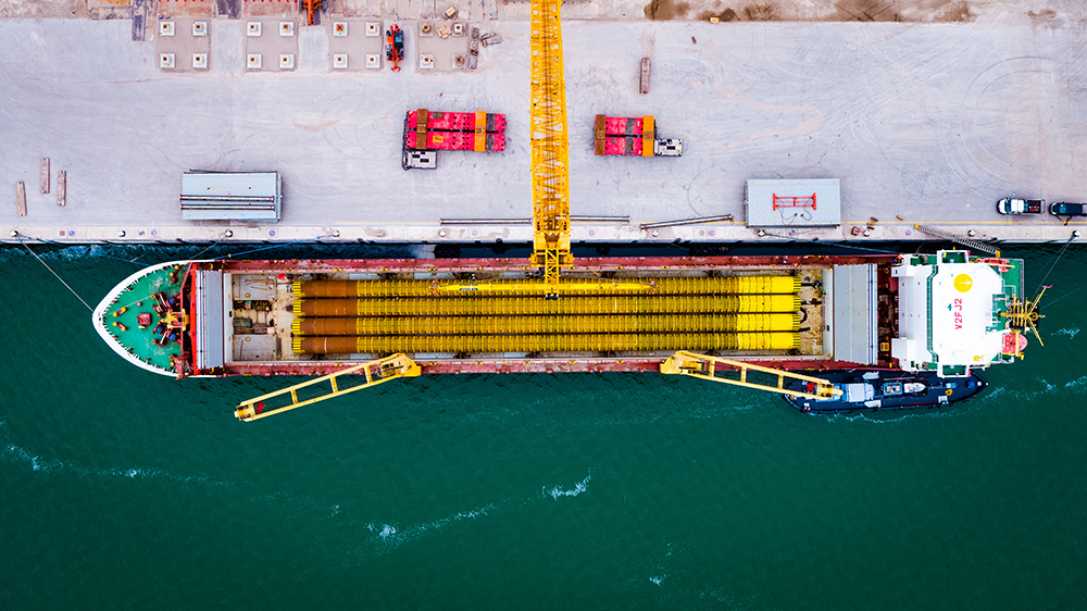 Aerial view of a large industrial ship docked at a port, loaded with long yellow pipes and equipped with cranes for loading and unloading.