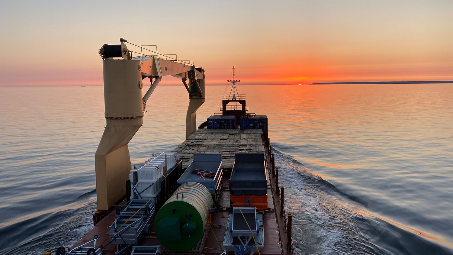 Cargo ship on the ocean at sunset, carrying containers and machinery, with a calm sea and colorful sky