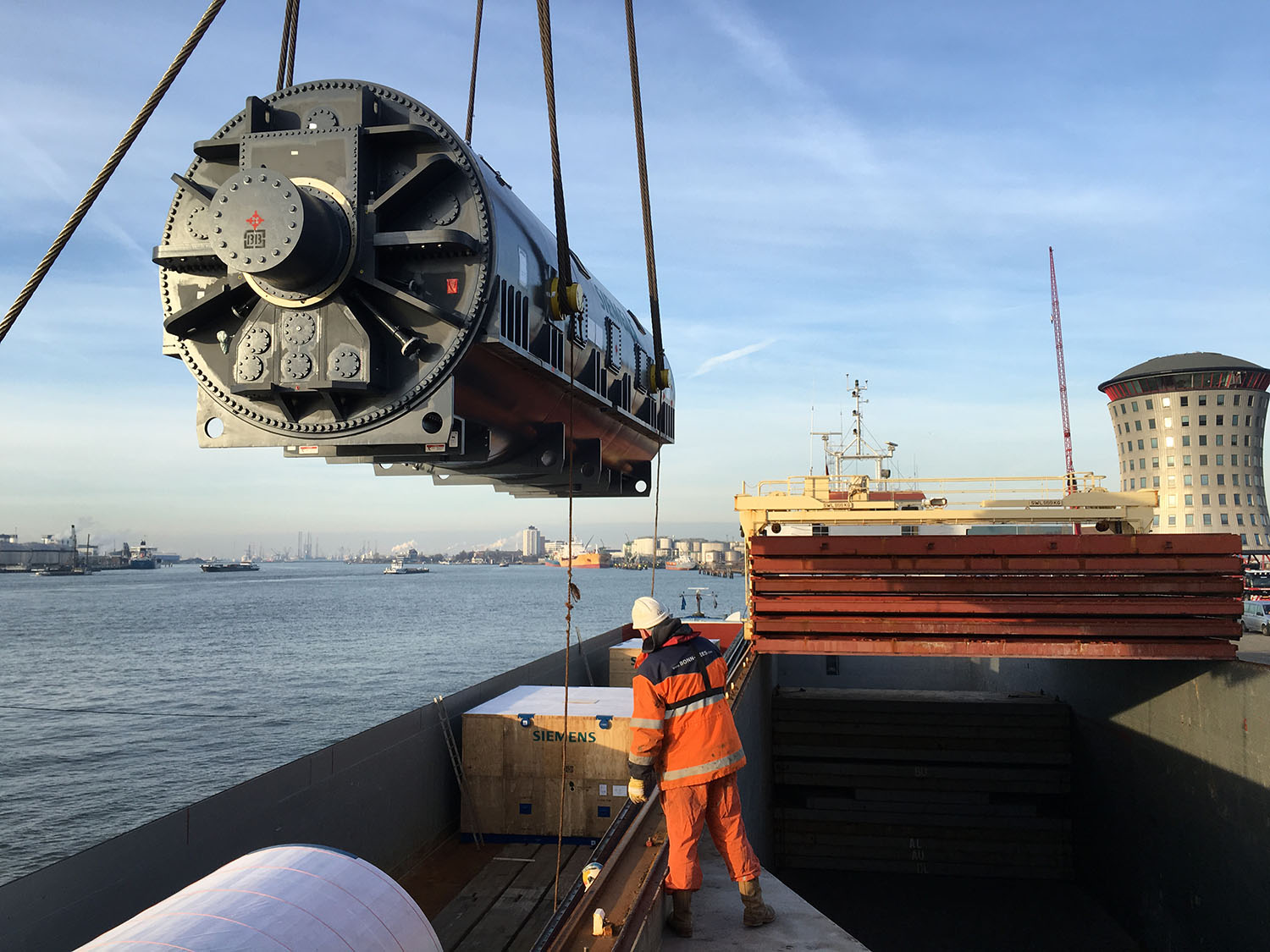 A large piece of industrial machinery is being lifted by a crane onto a cargo ship. A worker in high-visibility orange safety gear is guiding the process. The background shows a port with water, other ships, and a distinctively shaped building.
