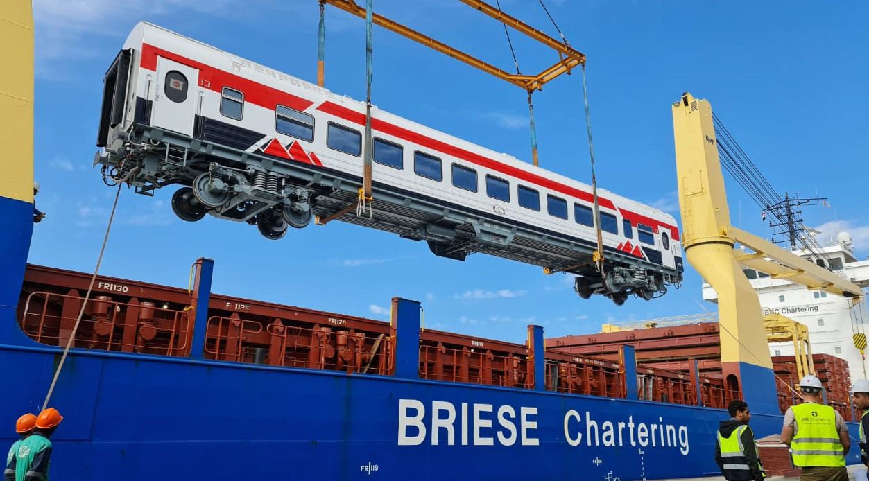 A powerful crane at a harbor carefully lifts a large train wagon and positions it onto a cargo ship.